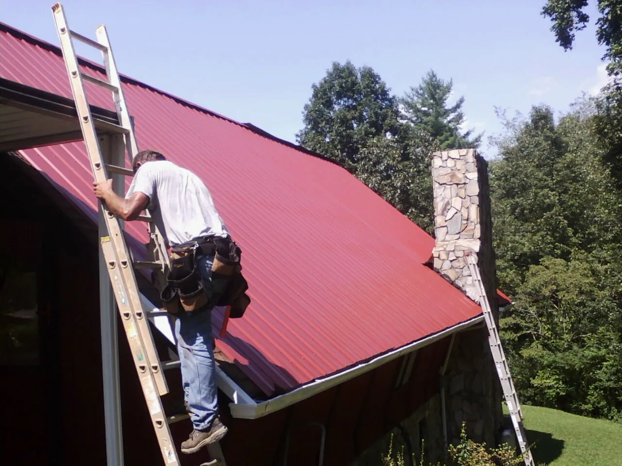 Roofer on a ladder beside a red metal roof and stone chimney — Soffit & Fascia Repair by Roofing Brothers in East Springfield