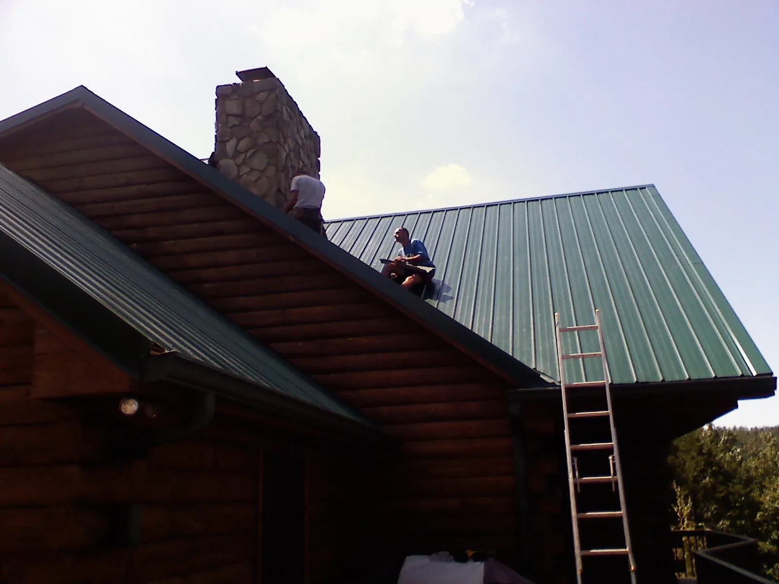 Roofers installing green metal panels beside a stone chimney on a log home in East Springfield